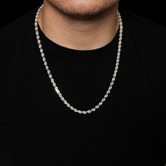 A close-up of a man wearing a silver-toned twisted rope chain necklace over a black t-shirt against a black background.